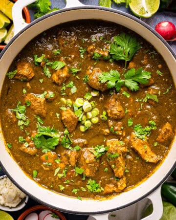 A pot of green chili stew (pork chile verde) with chunks of pork, topped with chopped green onions and cilantro, surrounded by limes, radishes, green onions, jalape&ntilde;os, and fresh herbs.