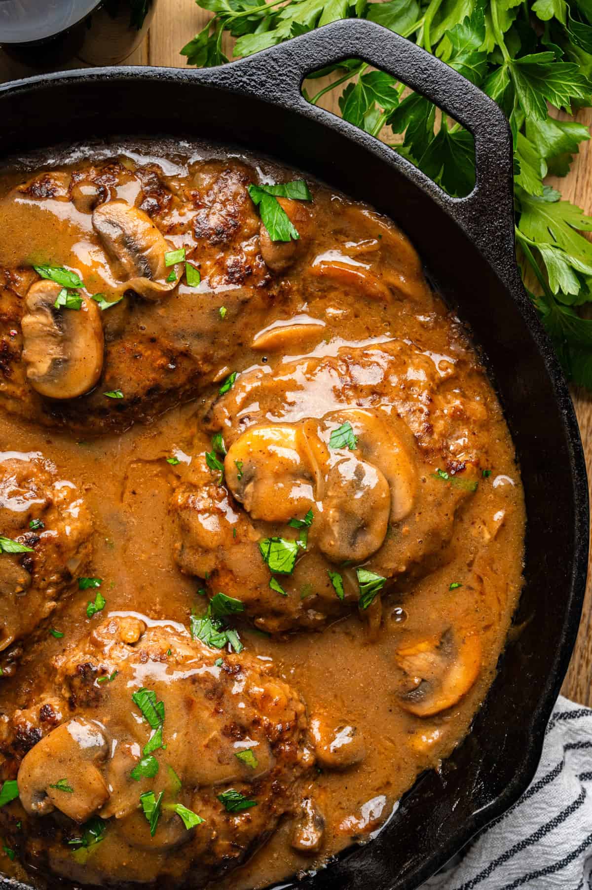Salisbury Steak with Brown Mushroom and Onion Gravy A cast-iron skillet filled with Salisbury steak patties topped with brown mushroom gravy, garnished with chopped parsley. Fresh parsley is visible in the background.