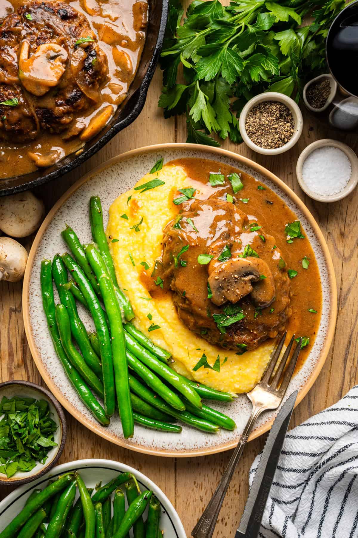 Salisbury Steak with Brown Mushroom and Onion Gravy. Serve with yellow polenta and green beans! A plate with Salisbury Steak patty in brown gravy over creamy polenta, garnished with parsley, and served with green beans. Surrounding the plate are herbs, seasonings, a skillet of meat, and a bowl of green beans.