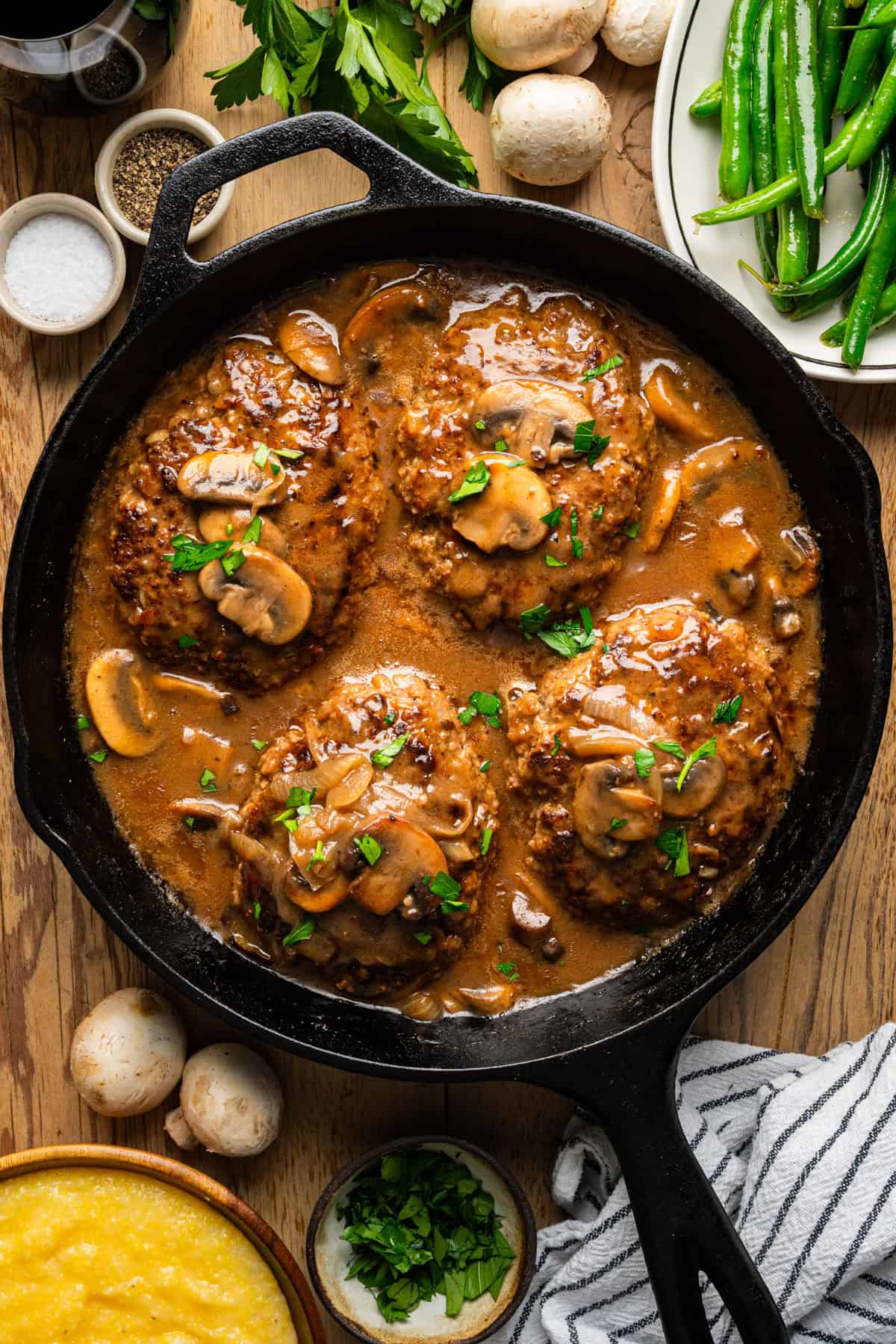 Salisbury Steak with Brown Mushroom and Onion Gravy A cast iron skillet filled with four Salisbury steaks in brown mushroom gravy, garnished with parsley. Surrounding the skillet are fresh green beans, mushrooms, seasonings, herbs, and a bowl of yellow polenta.