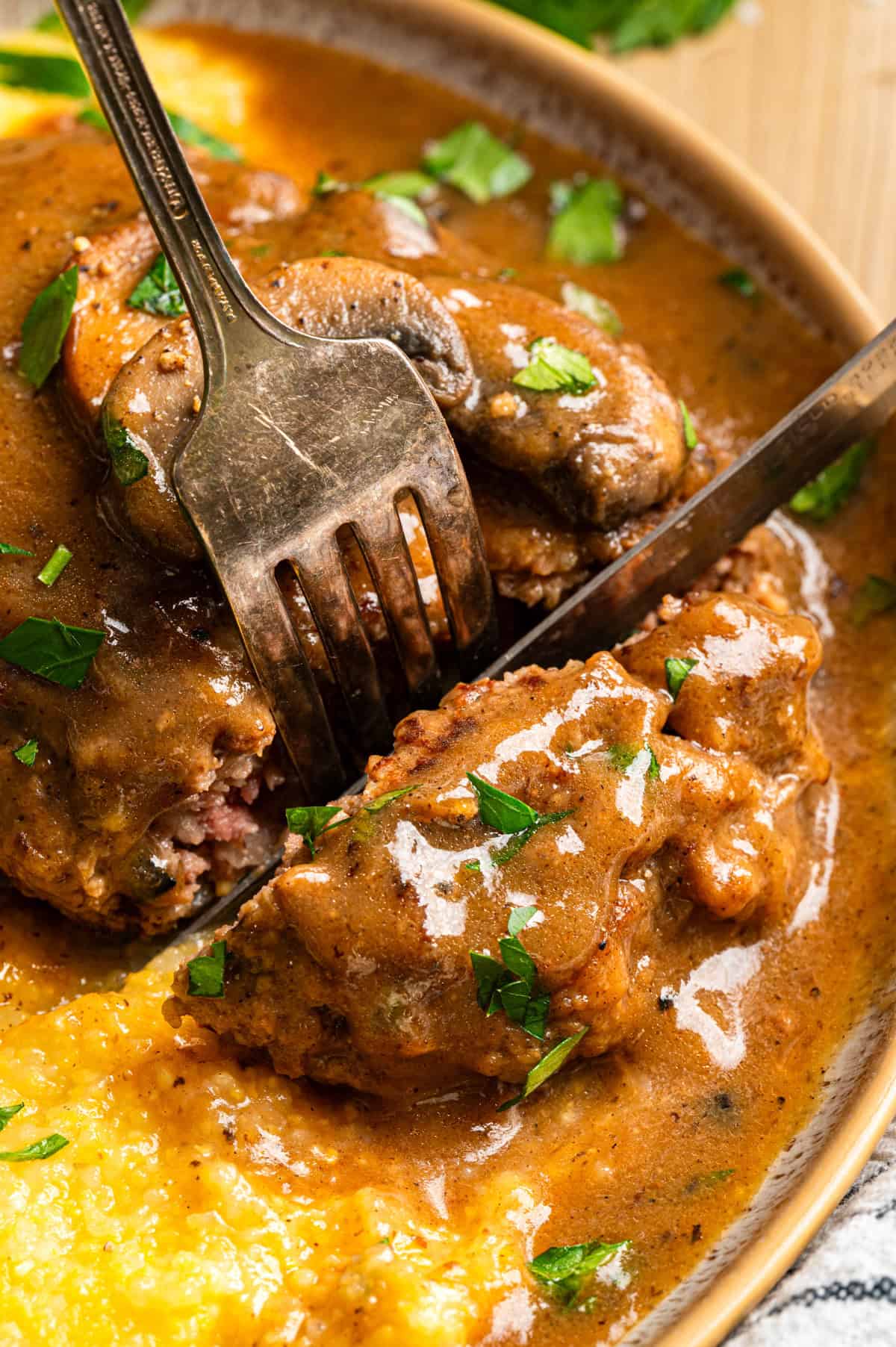 Salisbury Steak with Brown Mushroom and Onion Gravy. Serve with creamy polenta. A close-up of Salisbury steak covered in rich brown gravy, garnished with chopped parsley. A fork and knife are cutting into the steak, with creamy yellow polenta visible on the side.