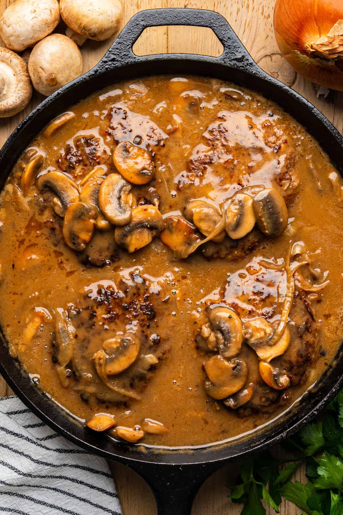 Salisbury Steak with Brown Mushroom and Onion Gravy A cast-iron skillet filled with Salisbury steaks cooked in rich brown mushroom gravy, topped with sliced mushrooms. Fresh mushrooms, an onion, parsley, and a striped towel are nearby on a wooden surface.