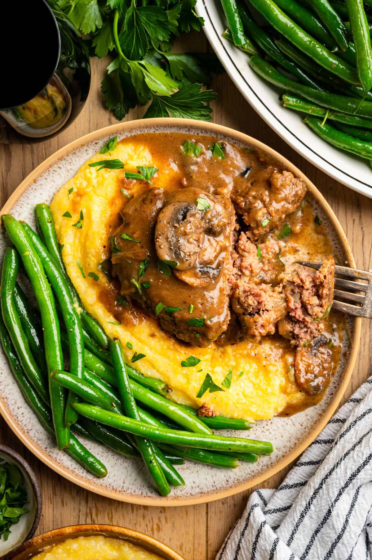 Salisbury Steak with Brown Mushroom and Onion Gravy A plate of Salisbury steak with mushroom gravy, served alongside creamy yellow polenta and fresh green beans, garnished with chopped parsley. A fork has a bite of steak, and extra green beans are nearby.