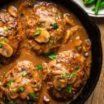 Four Salisbury steaks topped with sliced mushrooms and brown gravy, garnished with chopped parsley, served in a black cast iron skillet.