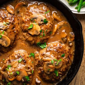 Four Salisbury steaks topped with sliced mushrooms and brown gravy, garnished with chopped parsley, served in a black cast iron skillet.