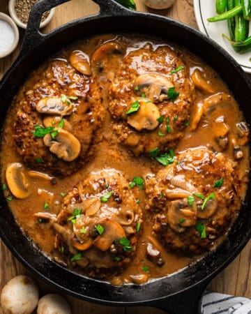 A cast iron skillet filled with four Salisbury steaks topped with mushroom gravy and chopped parsley, surrounded by fresh mushrooms, a plate of green beans, salt, pepper, and a striped napkin on a wooden table.