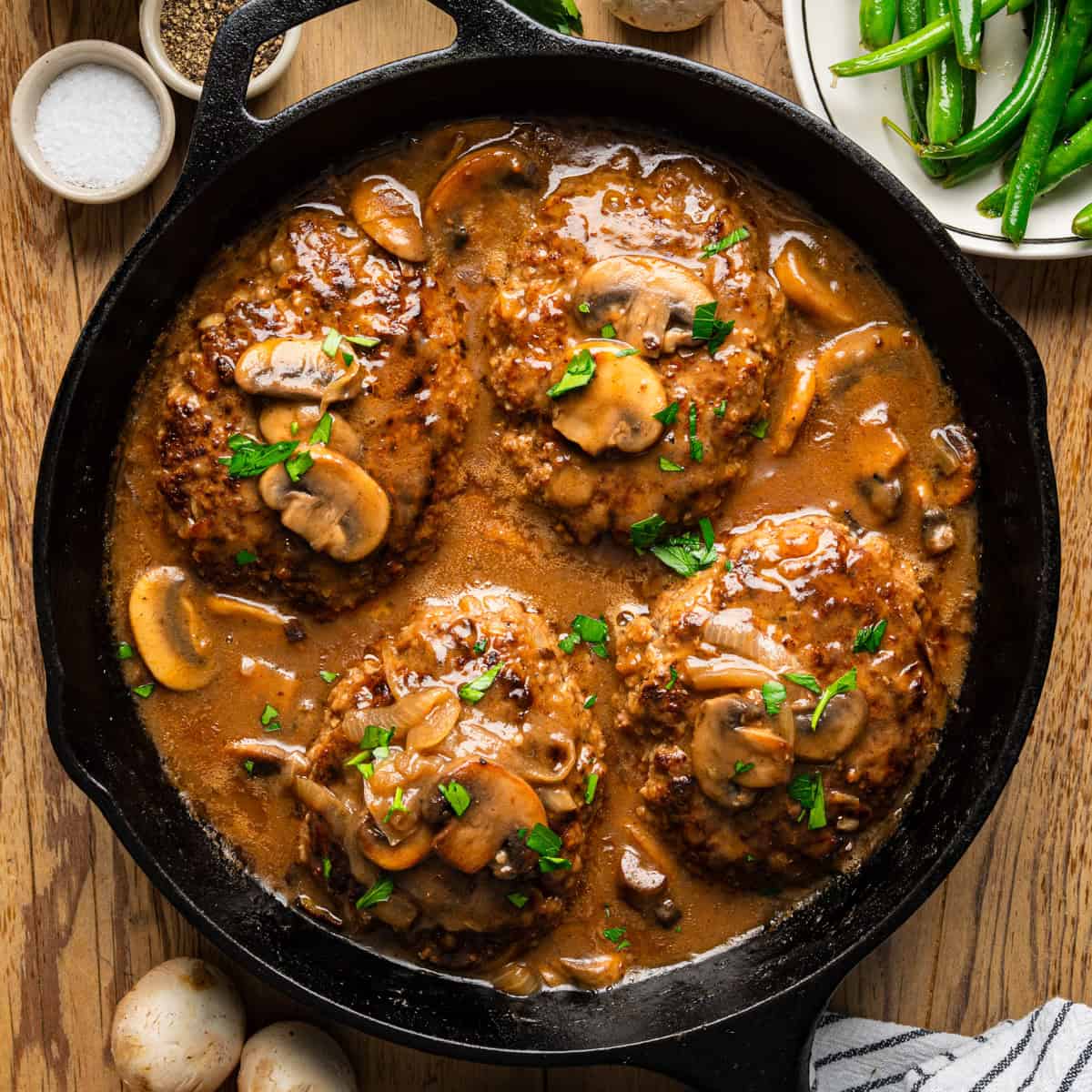 A cast iron skillet filled with four Salisbury steaks topped with mushroom gravy and chopped parsley, surrounded by fresh mushrooms, a plate of green beans, salt, pepper, and a striped napkin on a wooden table.