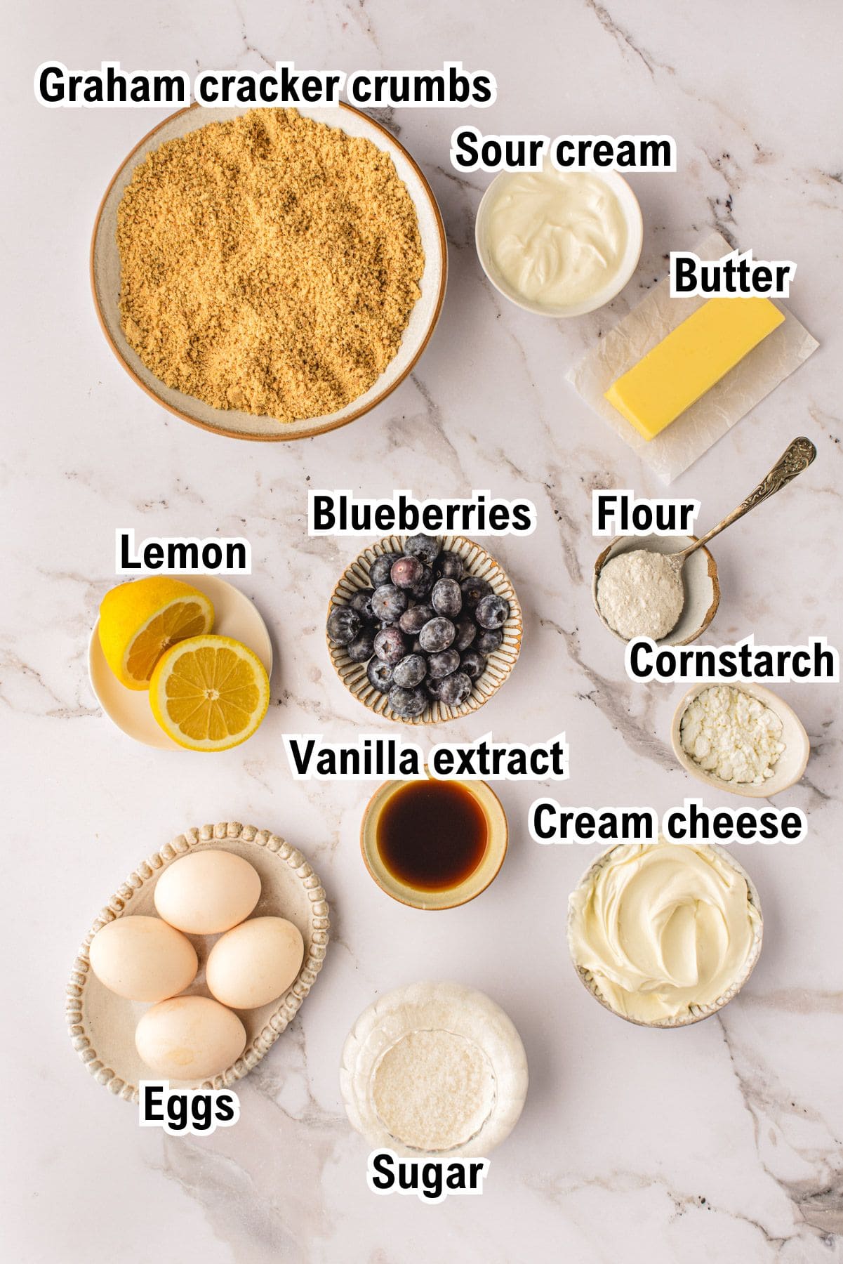 A flat lay of labeled blueberry cheesecake ingredients on a marble surface, including graham cracker crumbs, sour cream, butter, flour, cornstarch, cream cheese, sugar, eggs, vanilla extract, blueberries, and lemon slices.