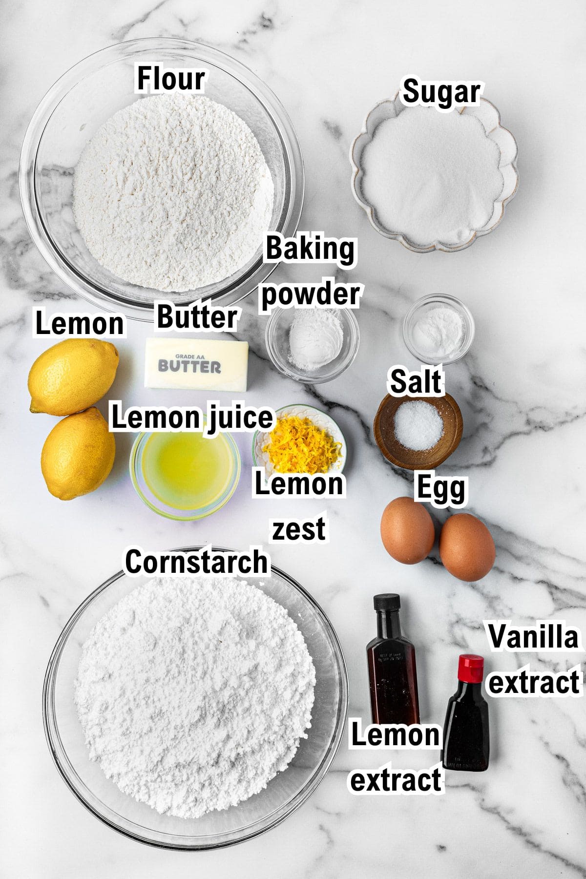 The Ingredients for Lemon Sugar Cookies This photo shows the ingredients for lemon sugar cookies: Two bowls with flour and cornstarch, a bowl of sugar, a stick of butter, two lemons, small bowls of lemon juice, lemon zest, and salt, an egg, baking powder, vanilla and lemon extract bottles on a white surface.