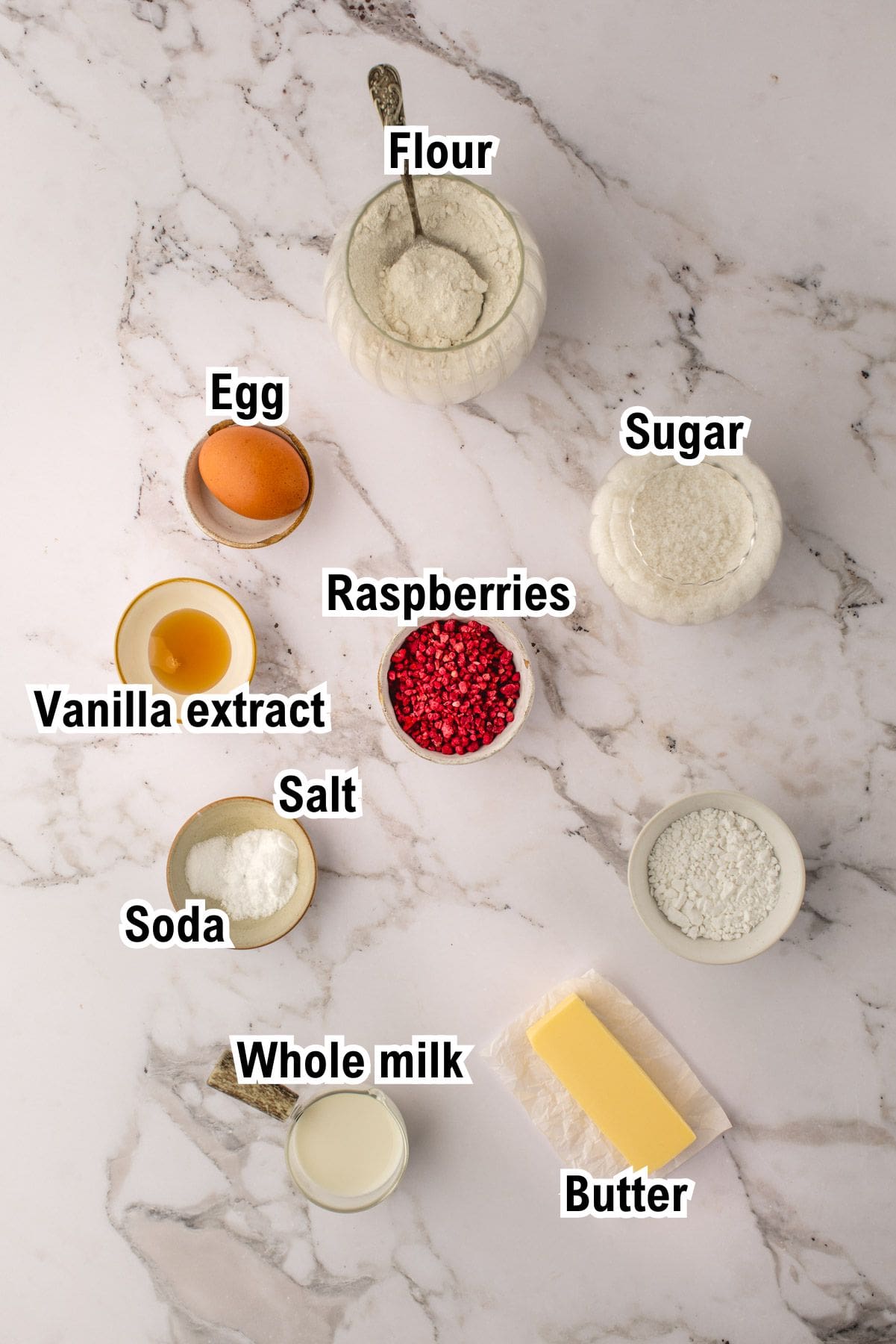 A flat lay of baking ingredients for Raspberry Cookies on a marble surface, labeled: flour, sugar, raspberries, egg, vanilla extract, salt, soda, whole milk, and a stick of butter. Each item is in a small bowl or container.