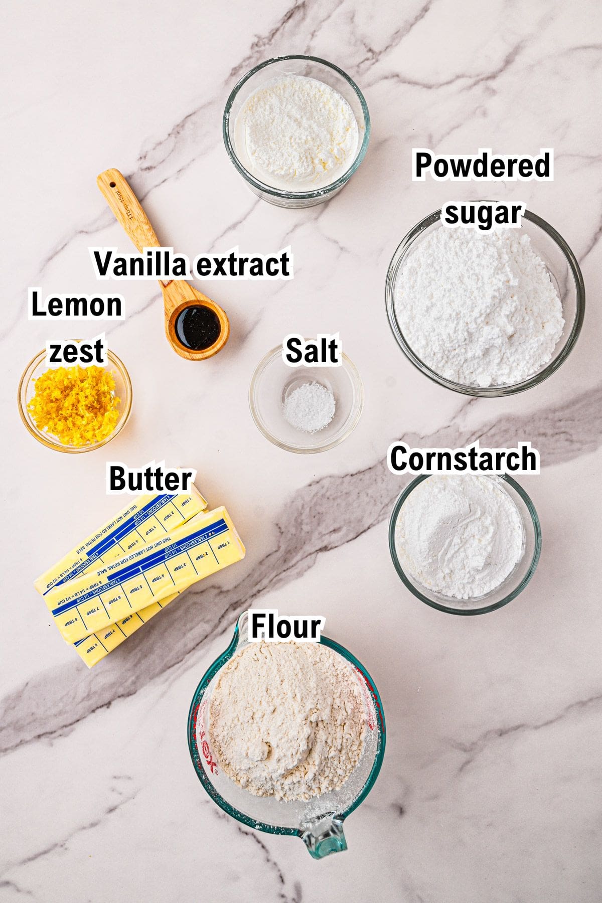 The Ingredients for Lemon Shortbread Cookies Measured ingredients for baking Lemon Shortbread Cookies are arranged on a marble surface: lemon zest, vanilla extract, powdered sugar, salt, cornstarch, butter, and flour, each labeled with text.