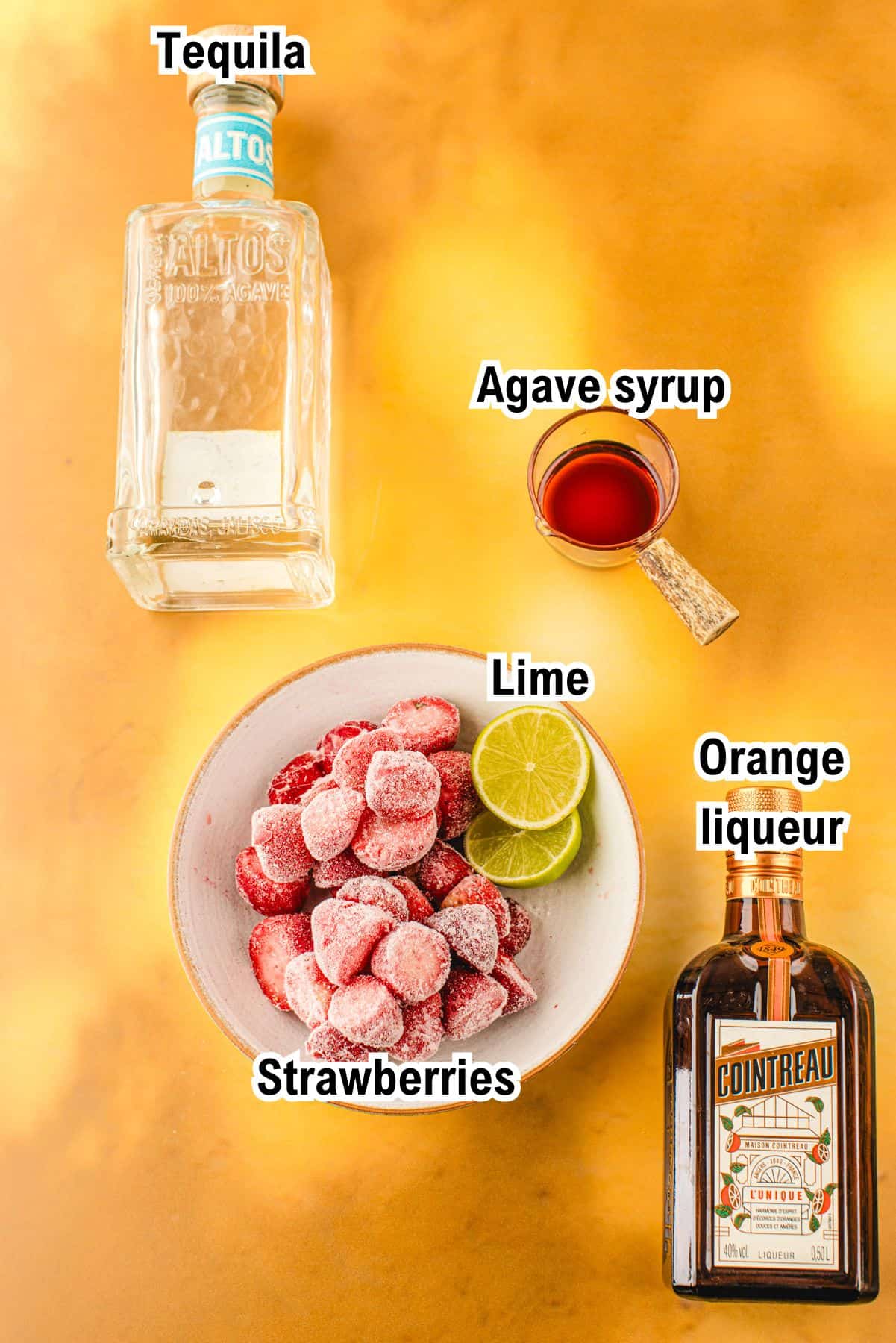 The Ingredients for Strawberry Margarita This photo shows the ingredients for Strawberry Margarita: Overhead view of ingredients for a cocktail on an orange surface: a tequila bottle, a bowl of frozen strawberries and lime slices, a small cup of agave syrup, and a bottle of orange liqueur. Labels identify each item.