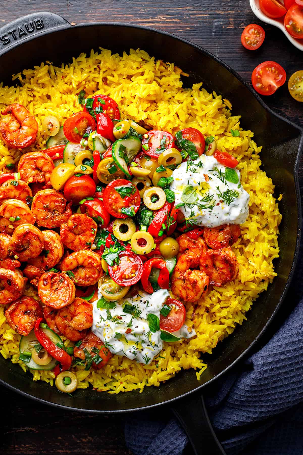 A skillet filled with yellow rice, spiced shrimp, sliced cherry tomatoes, cucumbers, olives, fresh herbs, and dollops of tzatziki, garnished with herbs and served on a dark background.