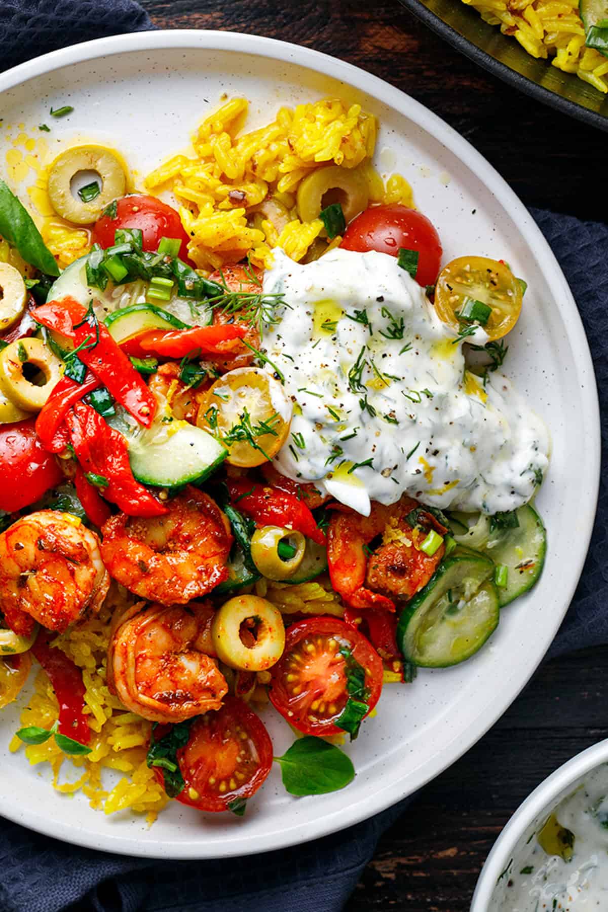 A plate of yellow rice topped with seasoned shrimp, sliced olives, cherry tomatoes, cucumbers, red peppers, fresh herbs, and a generous dollop of creamy tzatziki sauce.