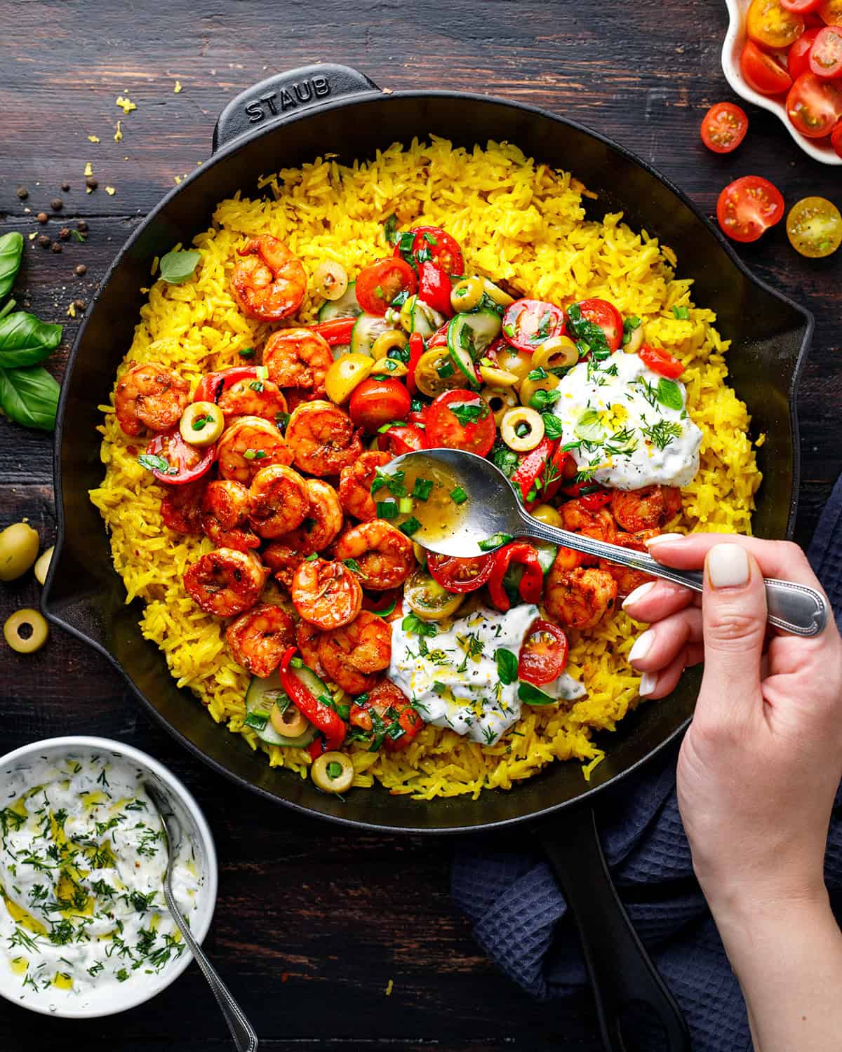 A skillet filled with yellow rice, spiced shrimp, sliced tomatoes, green olives, and dollops of tzatziki. A hand holds a spoon, ready to take a bite. Fresh ingredients and yogurt are nearby on a dark wooden table.