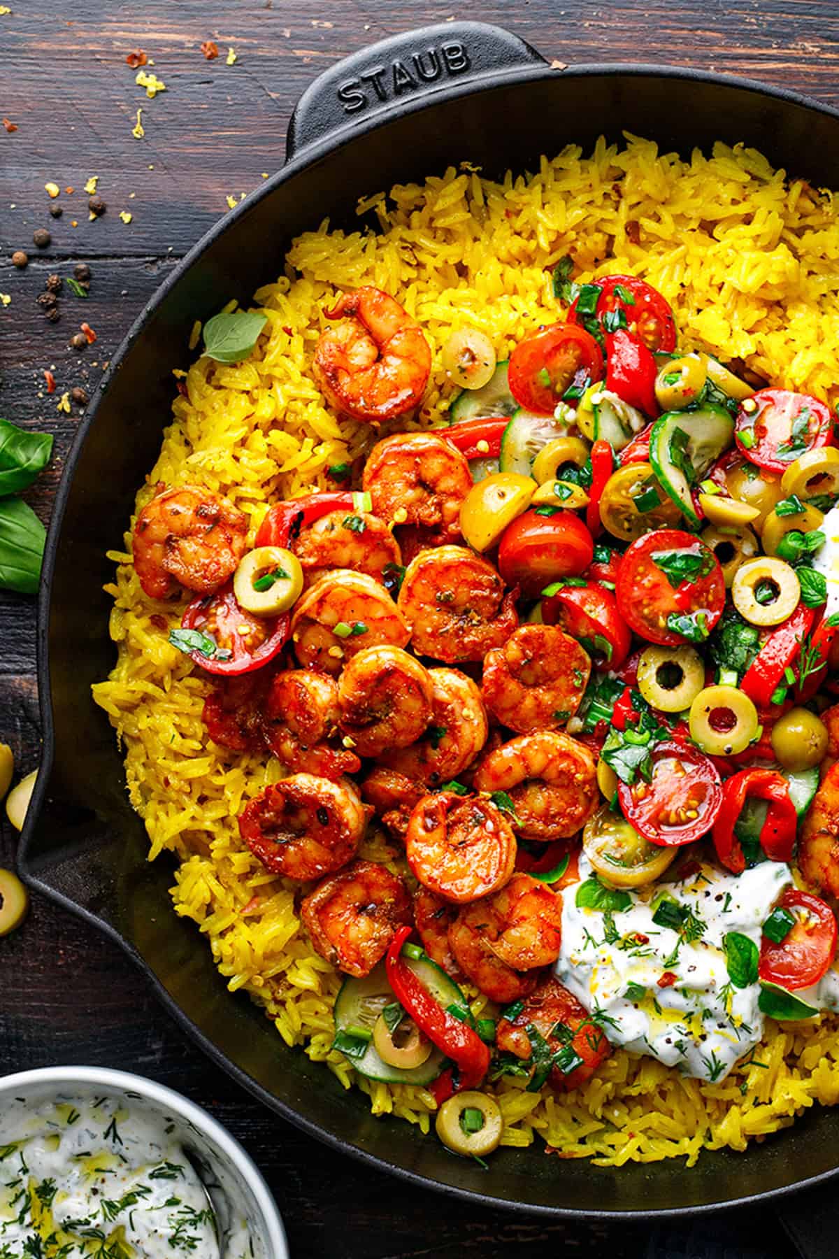 A cast iron skillet filled with yellow rice, seasoned shrimp, sliced green olives, cherry tomatoes, and a dollop of tzatziki, garnished with herbs on a rustic wooden table.