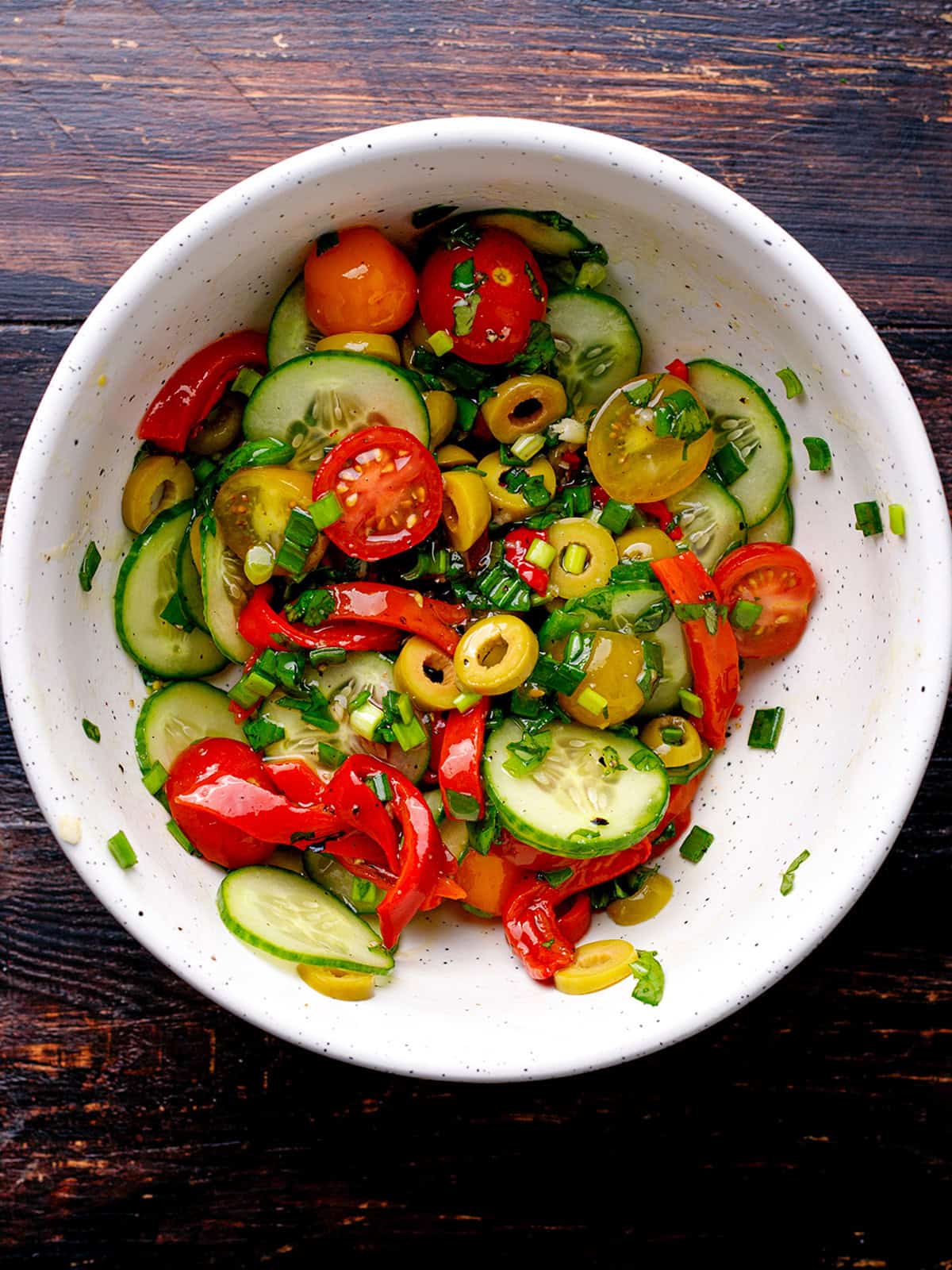 A white bowl filled with a colorful salad of sliced cucumbers, cherry tomatoes, red bell peppers, green olives, and chopped herbs on a dark wooden table.