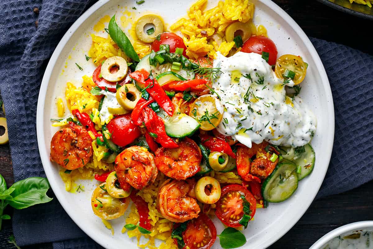 A plate of yellow rice topped with seasoned shrimp, sliced olives, cherry tomatoes, cucumbers, and herbs, served with a generous dollop of creamy tzatziki sauce on the side.