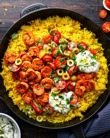 A skillet filled with yellow rice topped with spiced shrimp, halved cherry tomatoes, green olives, fresh herbs, and dollops of creamy tzatziki sauce, set on a dark wooden surface with extra tzatziki sauce and tomatoes nearby.