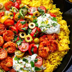 A vibrant dish with yellow rice, spiced shrimp, sliced tomatoes, green olives, cucumbers, fresh herbs, and generous dollops of creamy tzatziki sauce, all served in a black skillet.