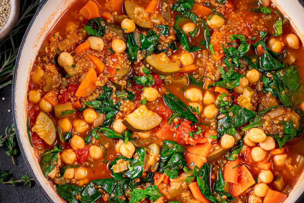 A close-up of a pot filled with colorful vegetable quinoa soup featuring chickpeas, spinach, carrots, zucchini, tomatoes, and quinoa simmered in a rich, red broth.