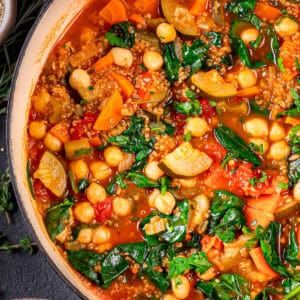 Close-up of a colorful vegetable quinoa soup with chickpeas, spinach, carrots, tomatoes, zucchini, and quinoa in a pot, garnished with fresh herbs.