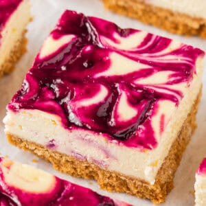 A square slice of blackberry cheesecake bar with a crumbly golden-brown crust and a creamy cheesecake layer, topped with a vibrant purple berry swirl pattern.