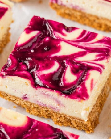 A close-up of a blackberry cheesecake bar with a graham cracker crust and a swirled raspberry sauce topping, showcasing creamy layers and vibrant purple and white patterns.