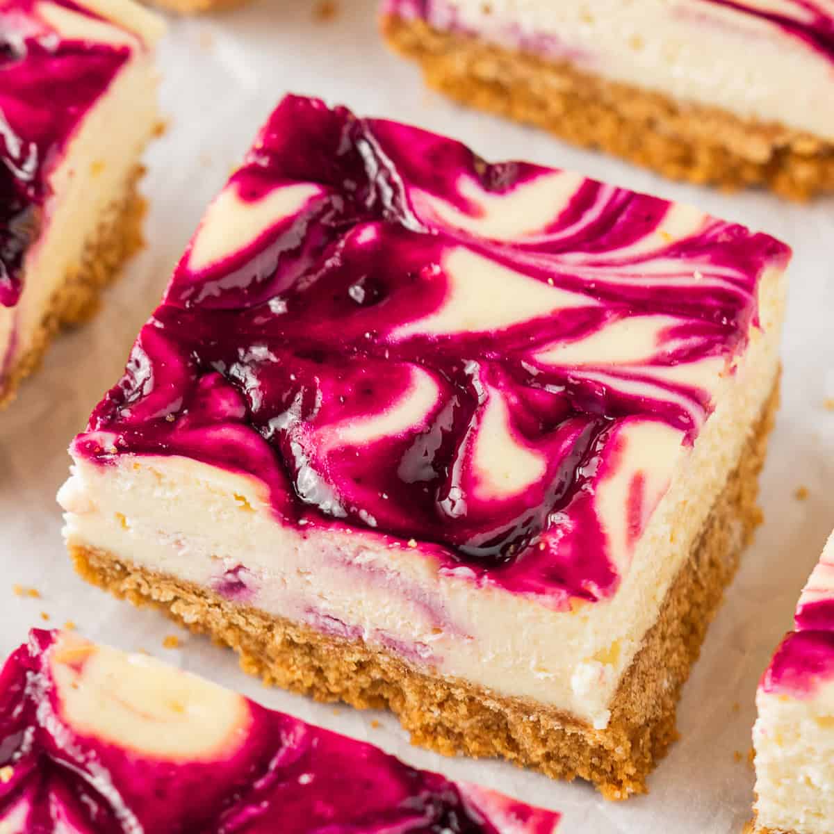 A close-up of a blackberry cheesecake bar with a graham cracker crust and a swirled raspberry sauce topping, showcasing creamy layers and vibrant purple and white patterns.