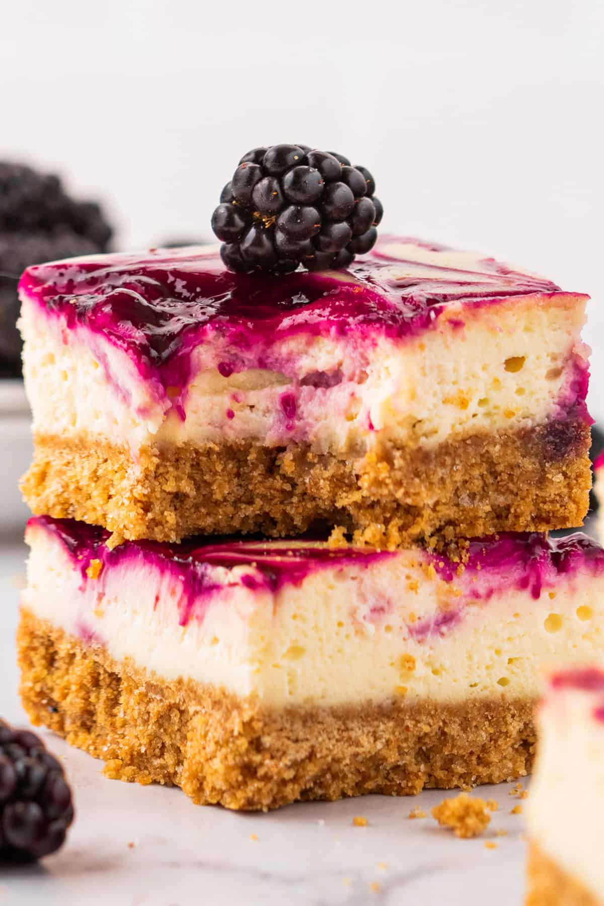 Two blackberry cheesecake bars with a graham cracker crust are stacked, topped with a layer of purple berry sauce and a fresh blackberry on top. 