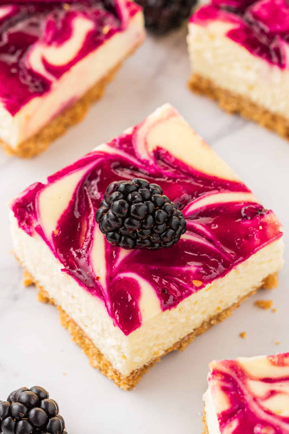 A square slice of blackberry cheesecake bar with a golden crust, topped with a vibrant blackberry swirl and a fresh blackberry, set on a white surface with other cheesecake bars nearby.