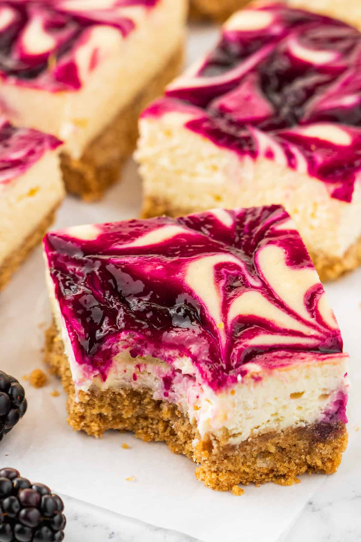 A square slice of blackberry cheesecake bar with a golden graham cracker crust and a vibrant purple blackberry swirl on top, with a bite taken out. Fresh blackberries are visible nearby.