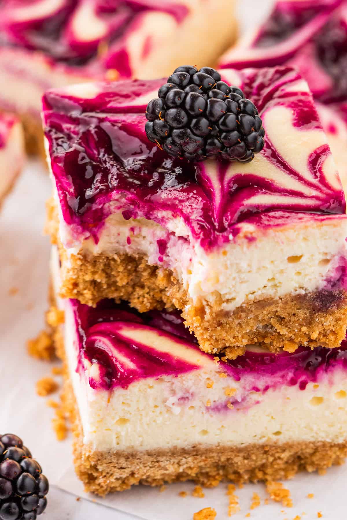 Two blackberry cheesecake bars with a golden graham cracker crust and vibrant purple berry swirl are stacked on top of each other. The top bar has a bite taken out and is garnished with a fresh blackberry.