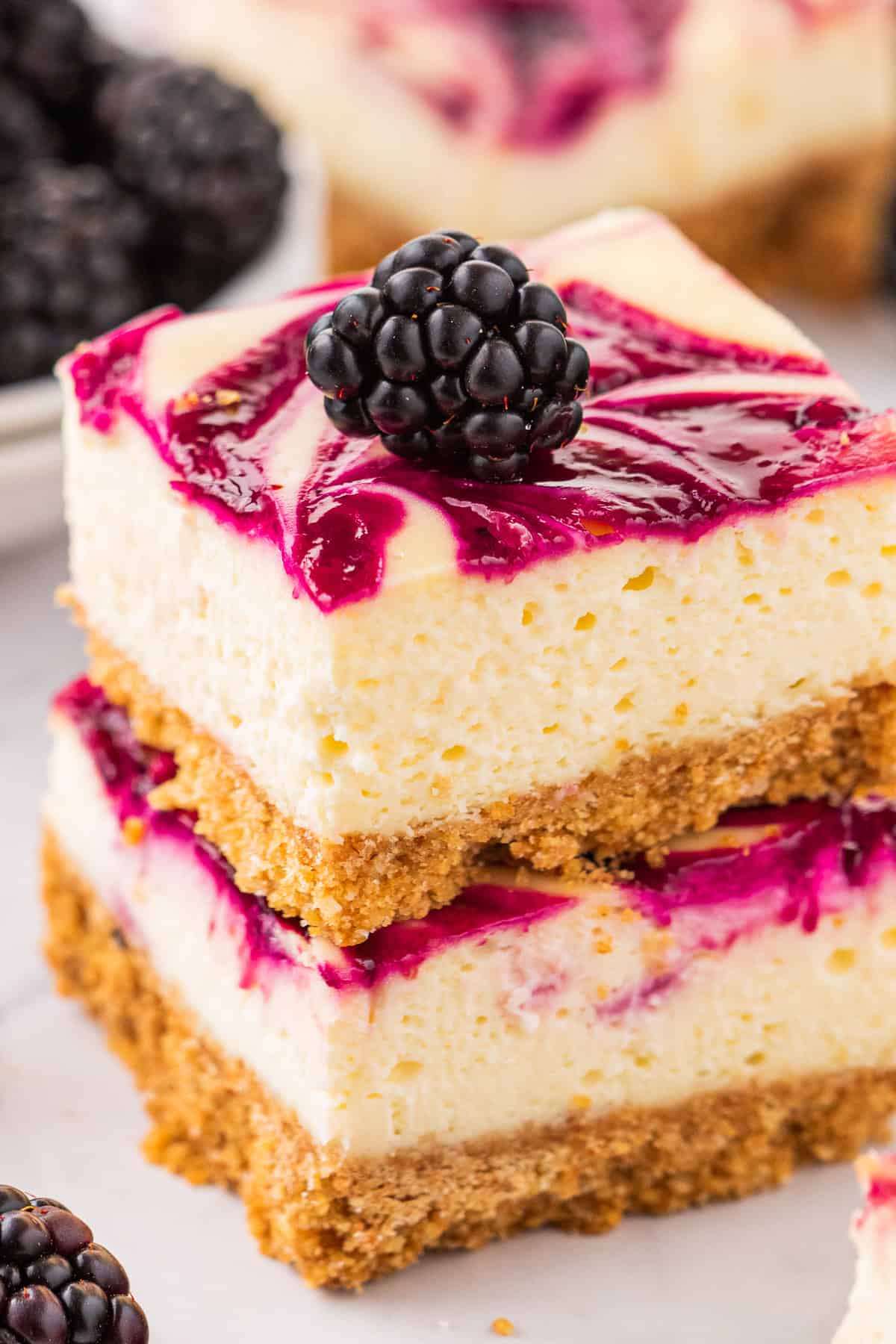 Two blackberry cheesecake bars with a golden graham cracker crust, topped with a swirl of purple berry sauce and a fresh blackberry, stacked on top of each other with more blackberries nearby.