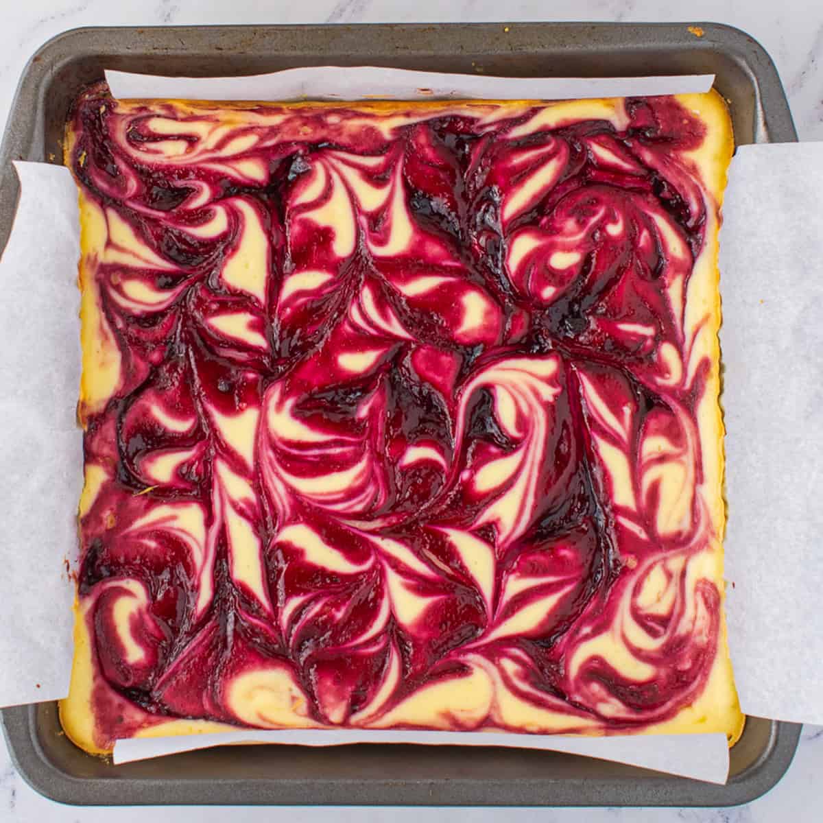 A square blackberry cheesecake in a baking pan with a vibrant red berry swirl pattern on top, set on white parchment paper.