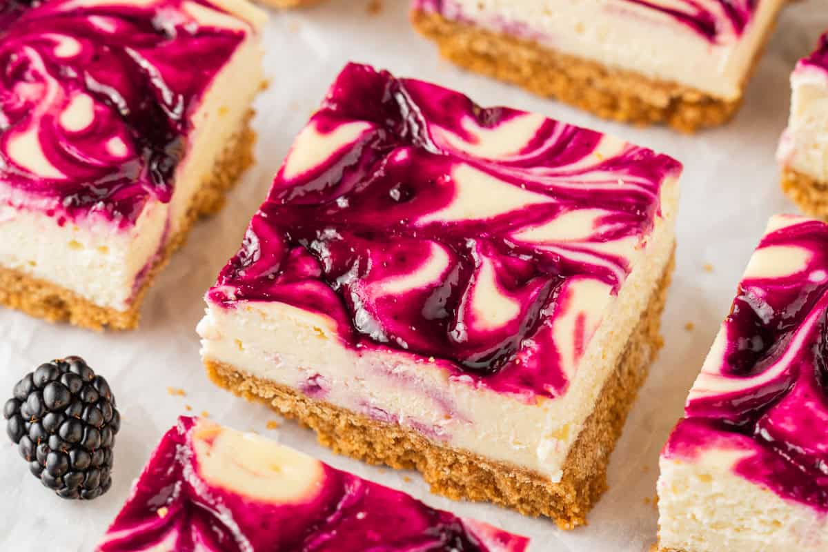 A close-up of blackberry cheesecake bars with a golden graham cracker crust and a swirled blackberry sauce topping, next to a fresh blackberry on a white surface.