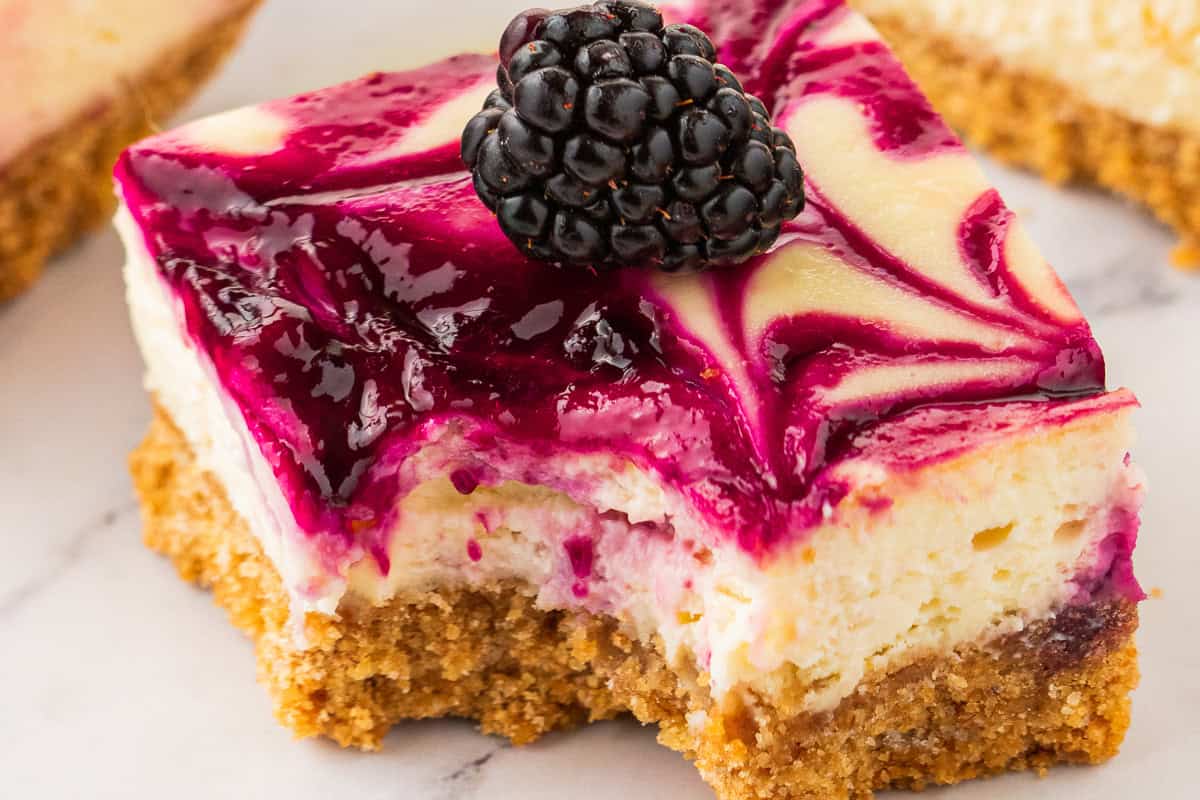 A blackberry cheesecake bar with a graham cracker crust, topped with a vibrant purple blackberry swirl and a fresh blackberry, has a bite taken out of it.