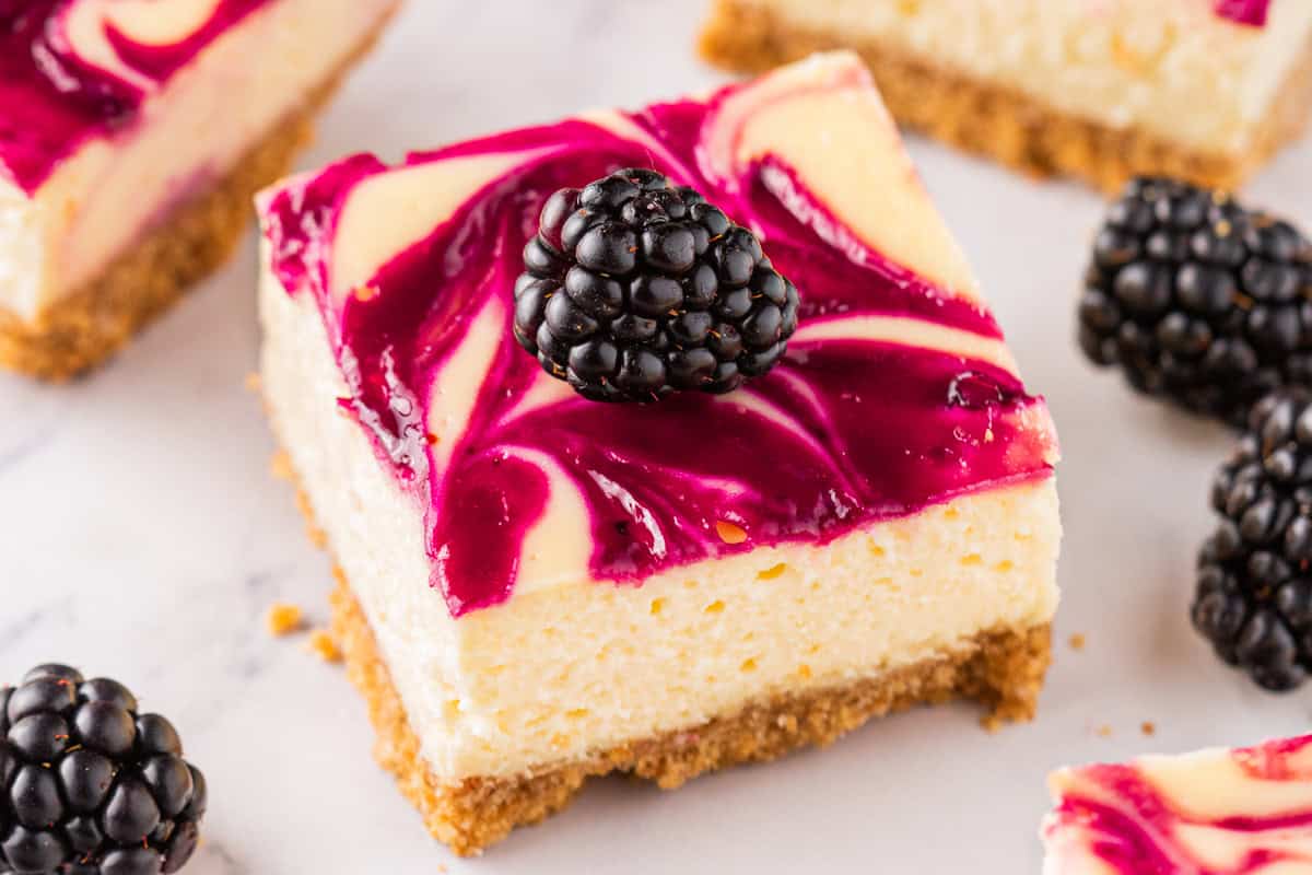A blackberry cheesecake bar with a graham cracker crust, topped with a swirl of purple berry sauce and a fresh blackberry, sits on a white surface with more blackberries scattered around.