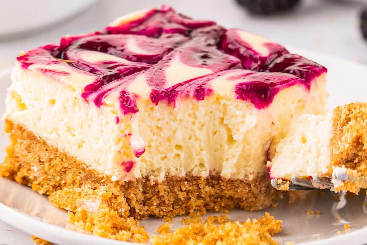 A close-up of a blackberry cheesecake bar with a crumbly graham cracker crust, creamy filling, and a vibrant purple berry swirl on top. A fork is cutting into the dessert, and crumbs are scattered on the plate.