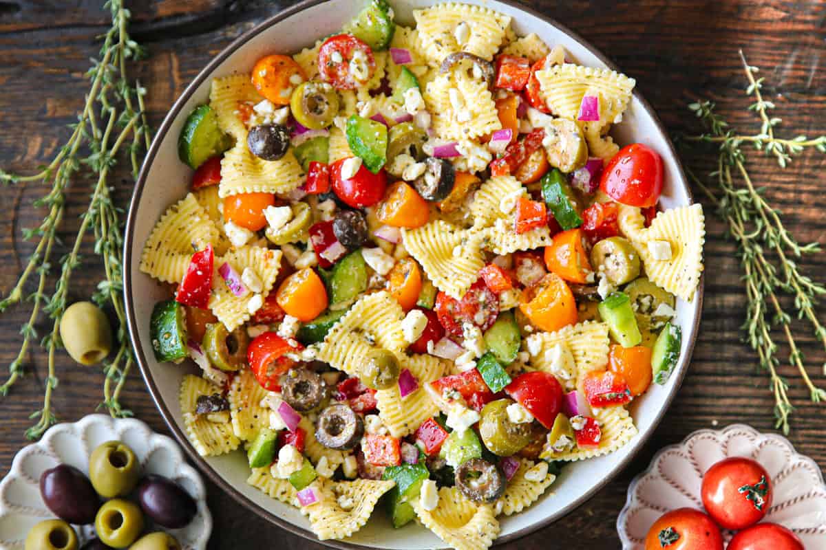 A bowl of Greek pasta salad with ridged bow tie noodles, cherry tomatoes, olives, green peppers, red onions, and feta cheese on a wooden table, garnished with fresh herbs. Small bowls of olives and tomatoes are nearby.