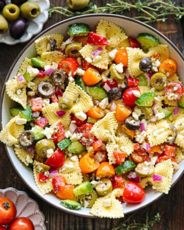 A bowl of Greek pasta salad with colorful cherry tomatoes, green and black olives, diced red onion, cucumber, feta cheese, and herbs on a wooden table, surrounded by small bowls of olives and tomatoes.