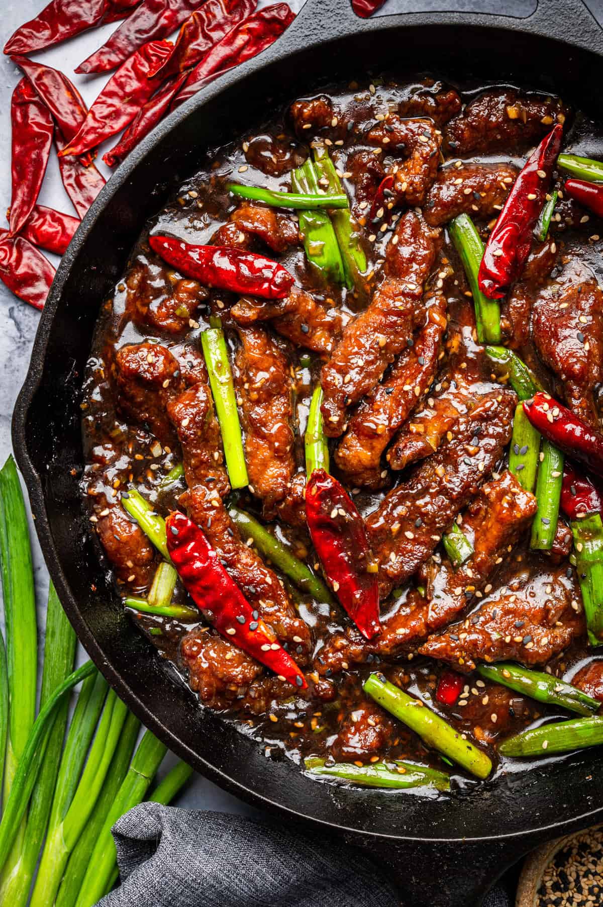 A skillet filled with Mongolian Beef: stir-fried beef strips, red chili peppers, and green onions in a glossy brown sauce, surrounded by dried red chilies and fresh green onions on the side.