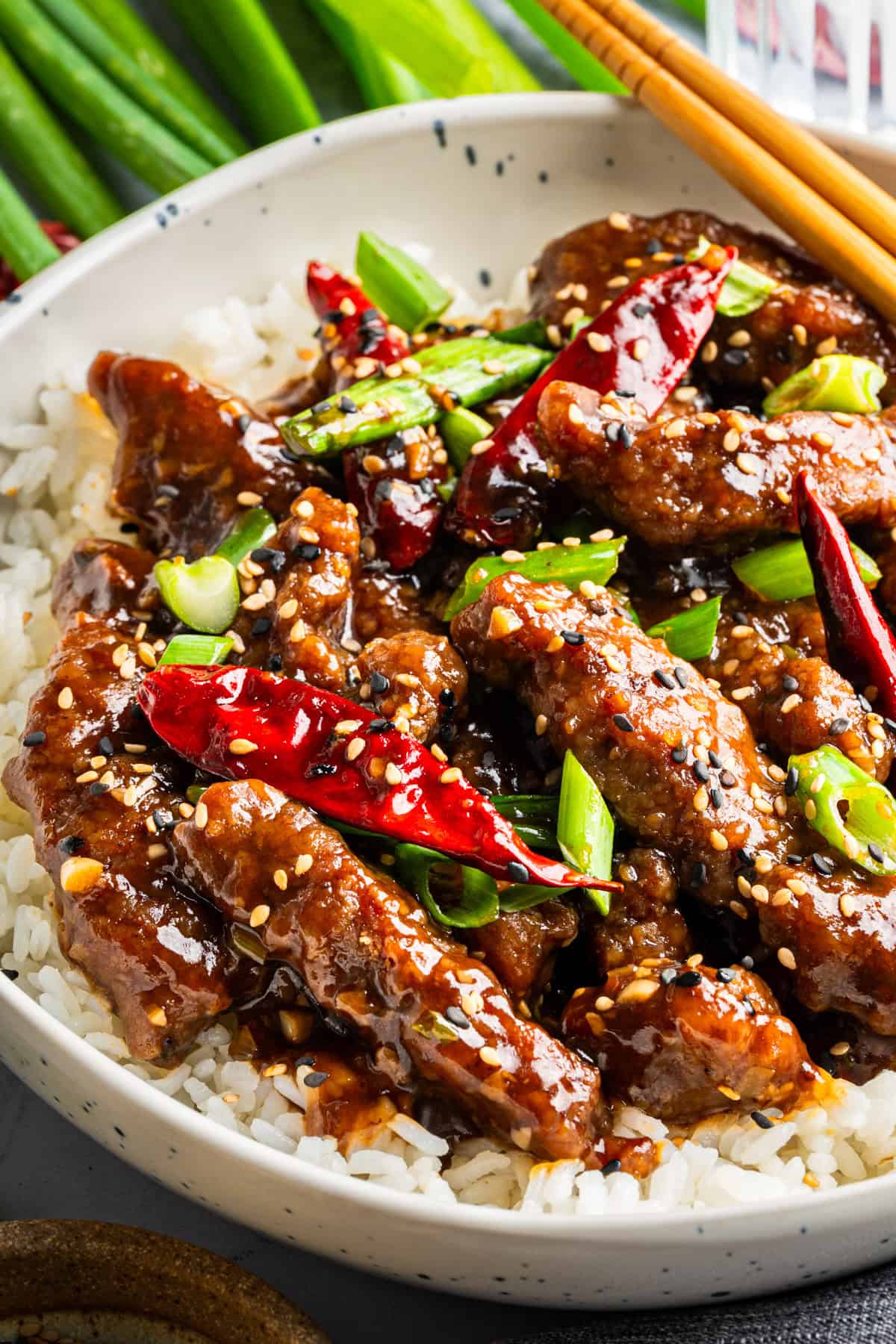 A bowl of white rice topped with crispy Mongolian beef in a dark, glossy sauce, garnished with red chili peppers, sesame seeds, and chopped green onions; chopsticks rest on the bowl's edge.