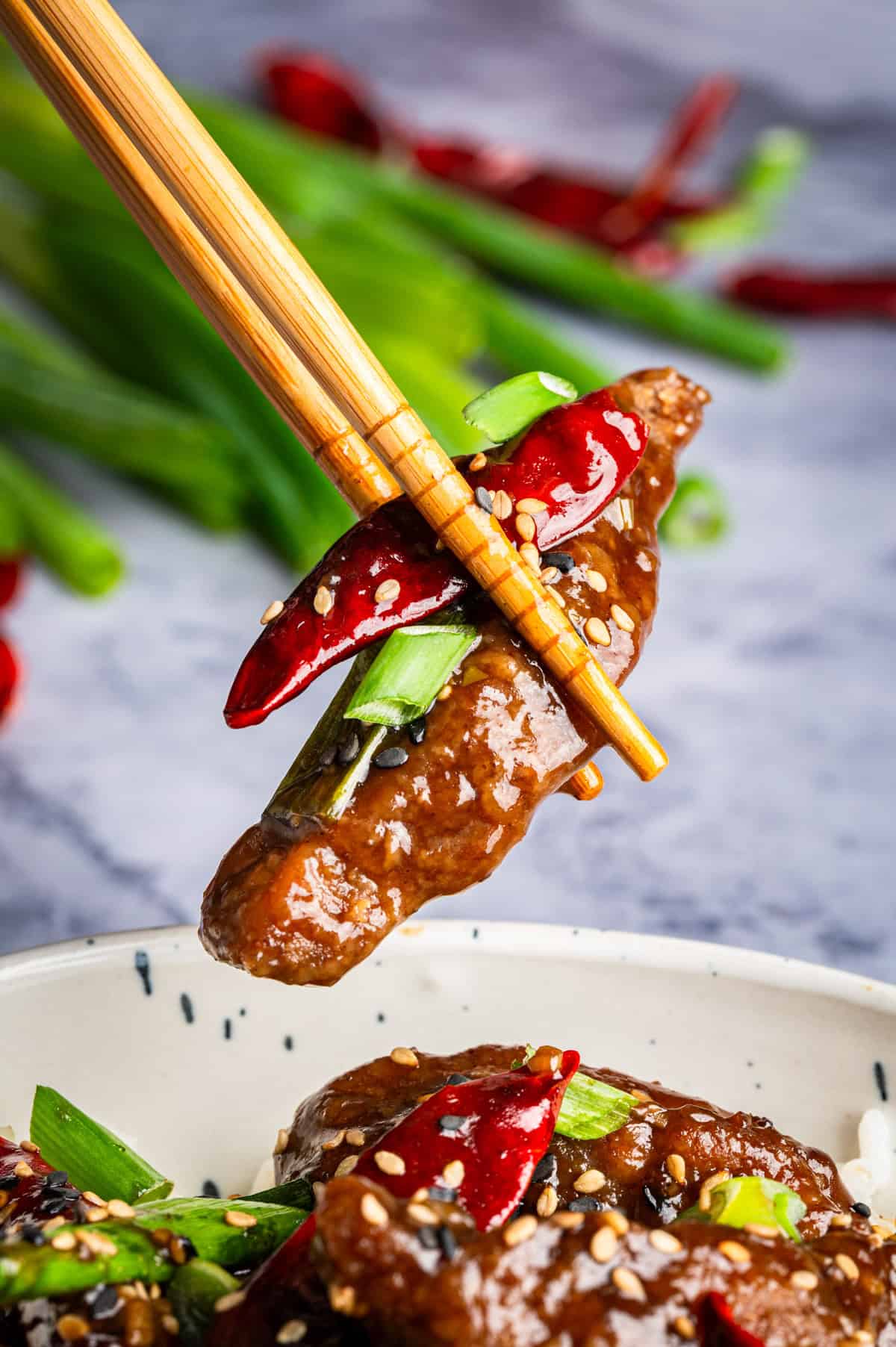 A pair of chopsticks holds a piece of glazed Mongolian beef garnished with sesame seeds, sliced green onions, and a red chili pepper. More beef, green onions, and peppers are visible in a bowl below.