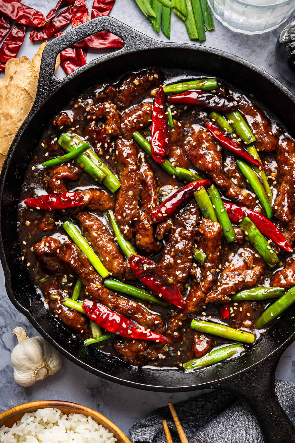 A skillet filled with stir-fried Mongolian beef strips in a glossy sauce, garnished with whole dried red chilies, green onions, and sesame seeds. Fresh ginger, garlic, and rice are visible nearby on a gray surface.