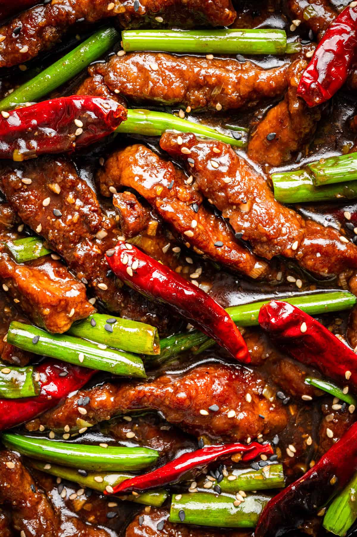 Close-up of stir-fried Mongolian beef strips with green onions, whole dried red chilies, and sesame seeds in a glossy, dark sauce. 