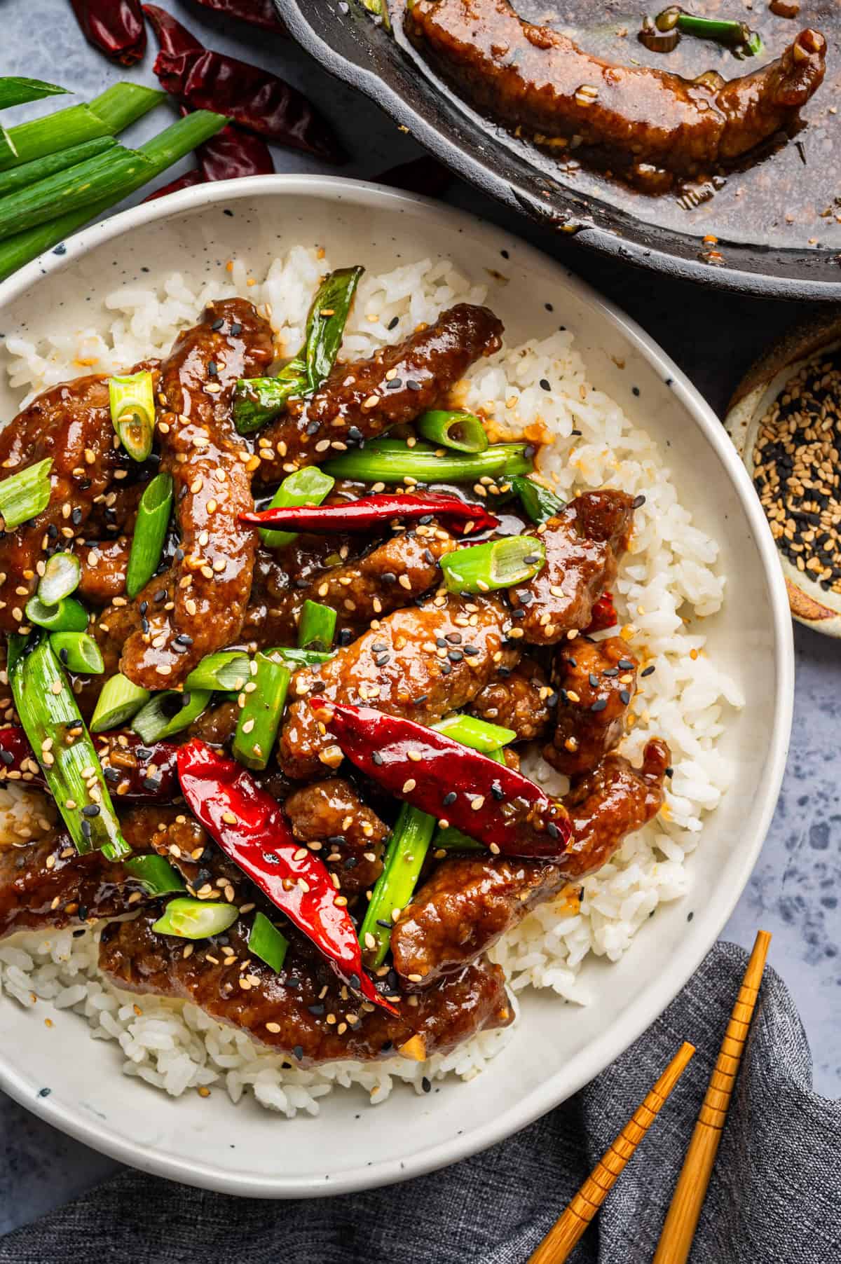 A bowl of white rice topped with saucy Mongolian beef strips, sliced green onions, whole dried red chilies, and sesame seeds, with chopsticks and a pan of beef in the background.