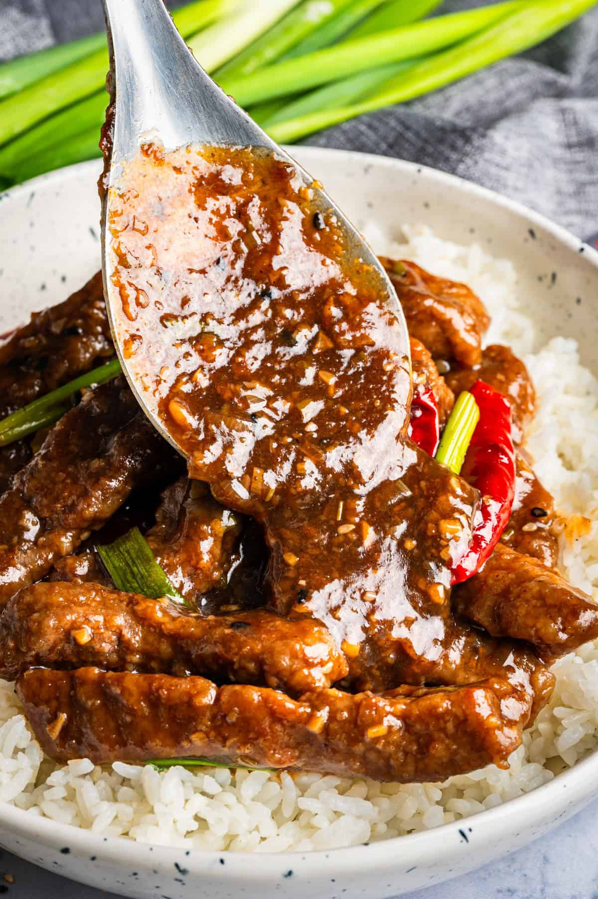 A close-up of a spoon pouring rich brown sauce over Mongolian beef strips and white rice in a bowl, garnished with green onions and dried red chili peppers.
