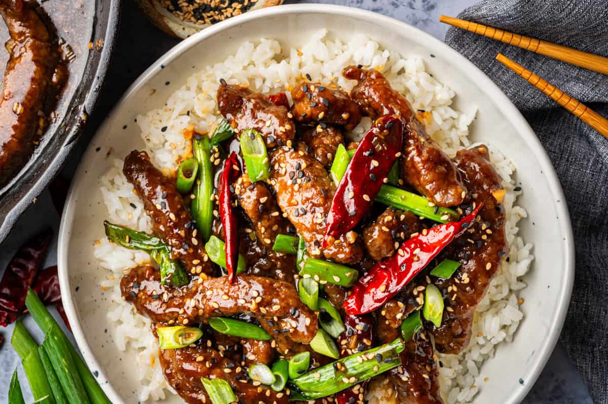 A bowl of white rice topped with crispy Mongolian beef strips, sliced green onions, dried red chilies, and sesame seeds, with chopsticks and a gray napkin beside the bowl.