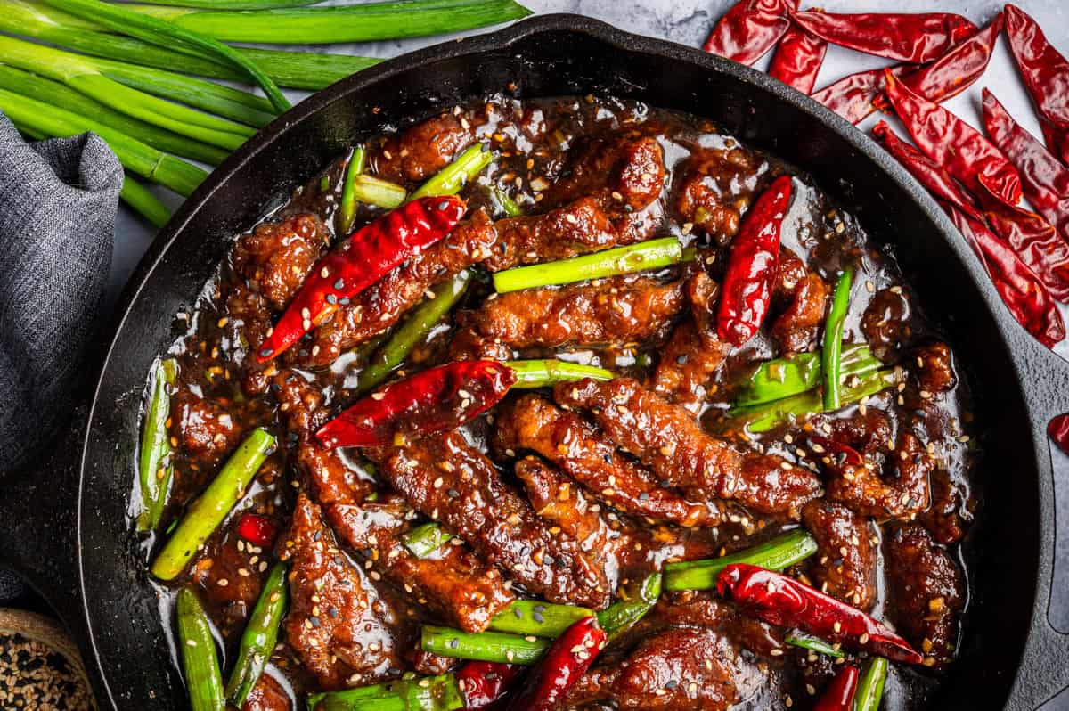 A skillet filled with saucy Mongolian beef strips, whole dried red chilies, and green onions, garnished with sesame seeds. Fresh green onions and dried chilies are scattered around the pan.